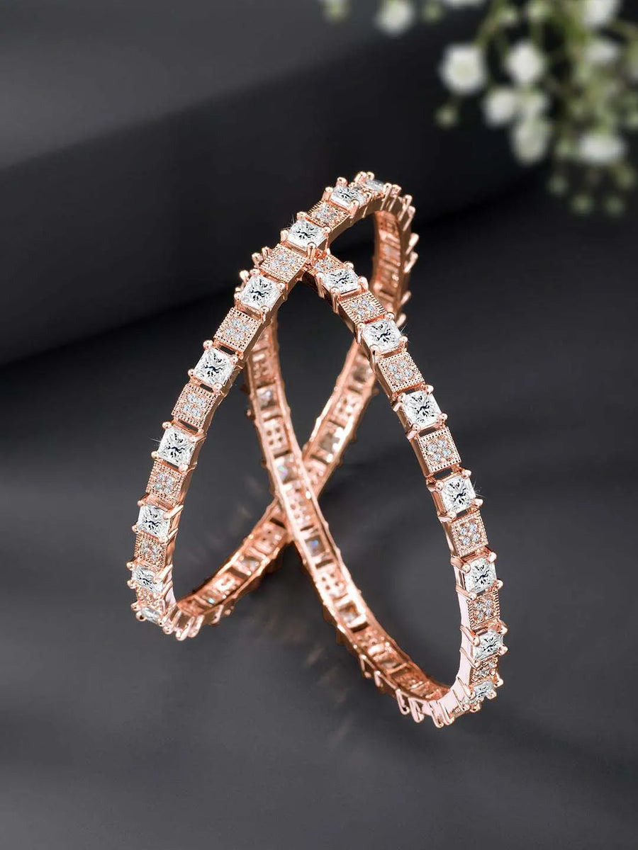 A set of two rose gold-colored bangles adorned with cubic zirconia stones, displayed against a dark background with floral patterns.