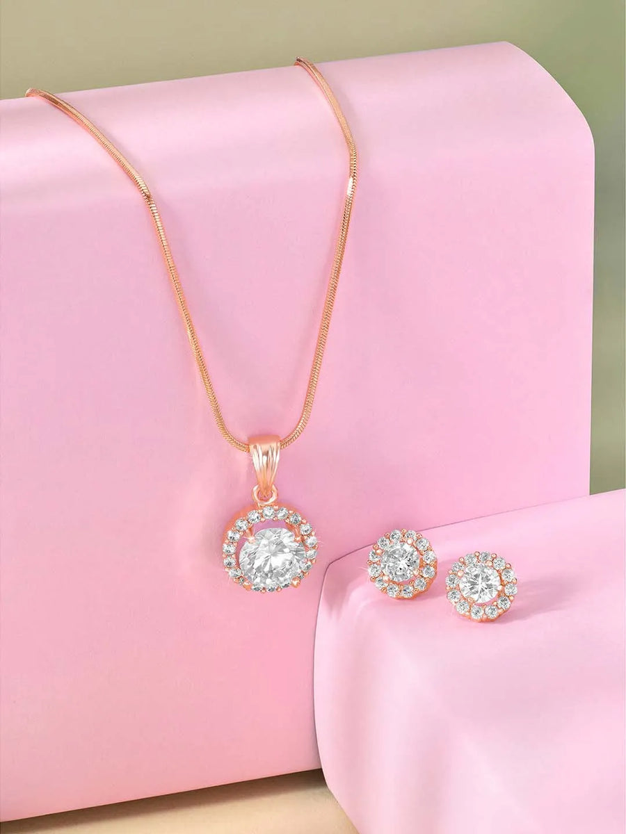 A rose gold-colored pendant necklace with a round cubic zirconia solitaire pendant accompanied by a pair of matching earrings, displayed on a pink cushion.