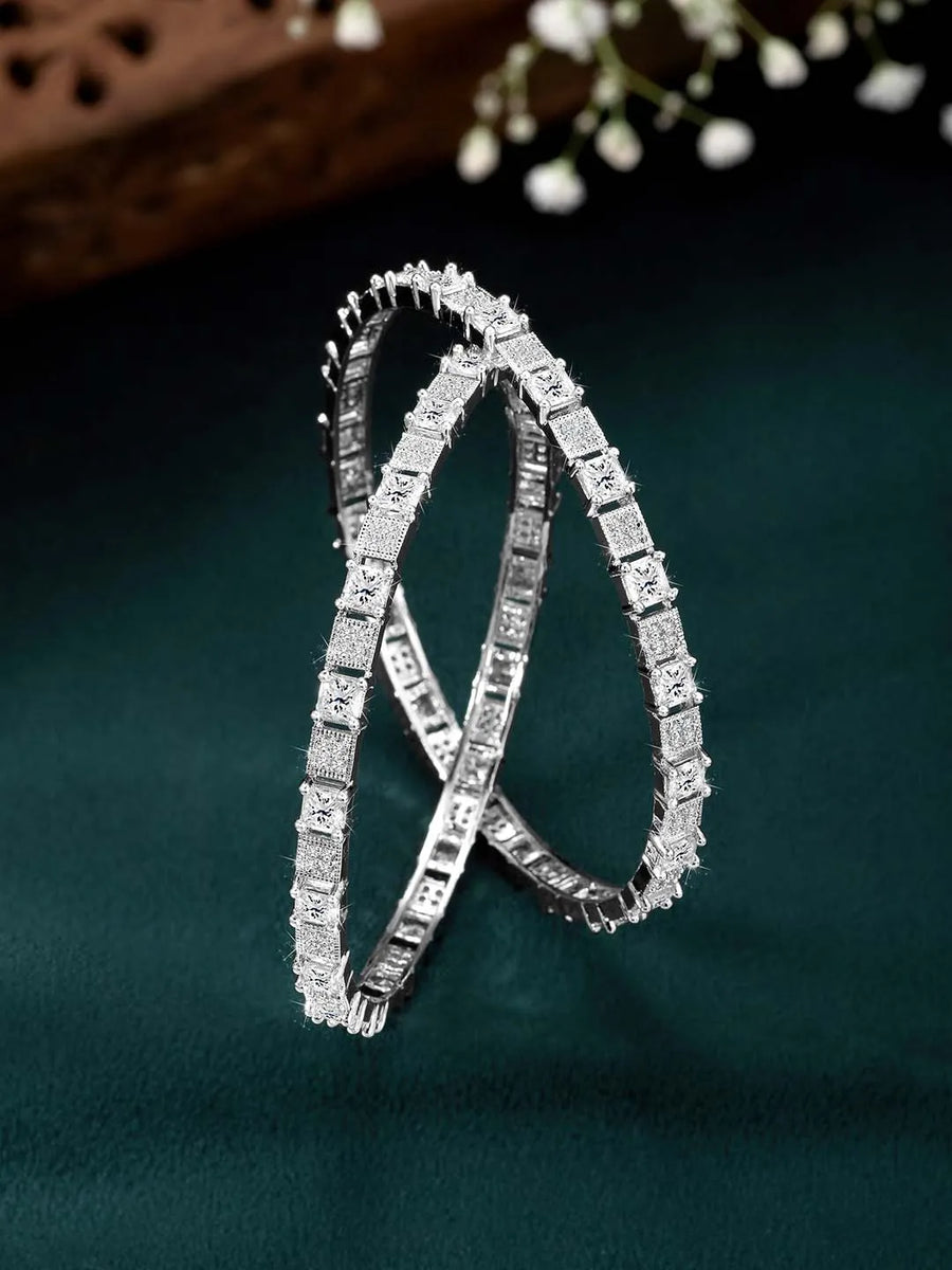 A set of two silver-colored bangles adorned with cubic zirconia stones, displayed against a dark background with floral patterns.